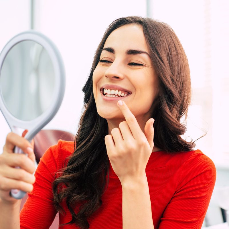 Brilliant work. Pretty girl in dentist cabinet is using a mirror to look through the ideal work of a doctor. She has a special dentist napkin over her vibrant red sweater