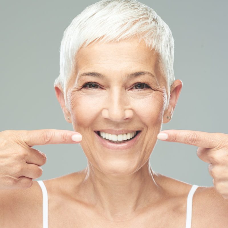 Beautiful caucasian smiling senior woman with short grey hair pointing at her teeth and looking at camera. Beauty photography.