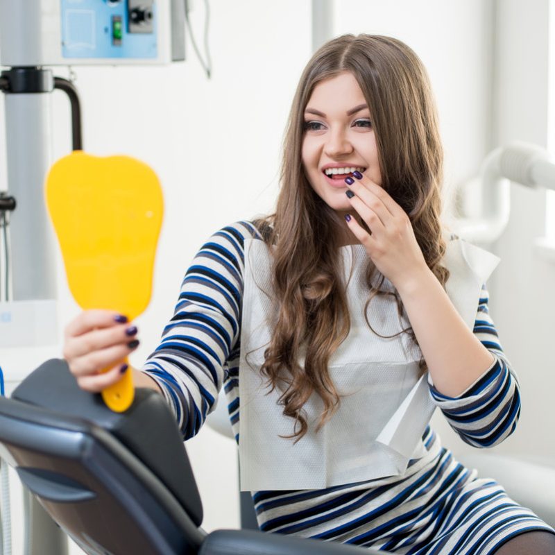 Beautiful young female patient with perfect white teeth sitting in dental chair, looking in the mirror and checking up teeth after treatment at modern dental clinic. Dentistry and healthcare concept