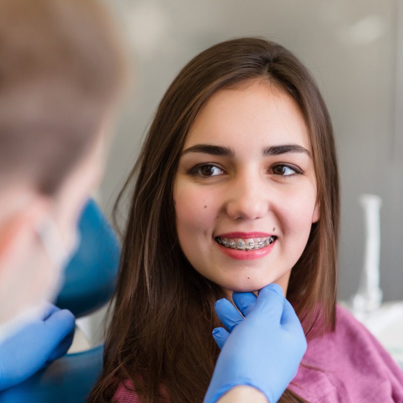 Close up of woman with brackets receiving dental braces treatment in clinic. Orthodontist using dental mirror and forceps while putting orthodontic braces on patient teeth. Concept of dentistry.