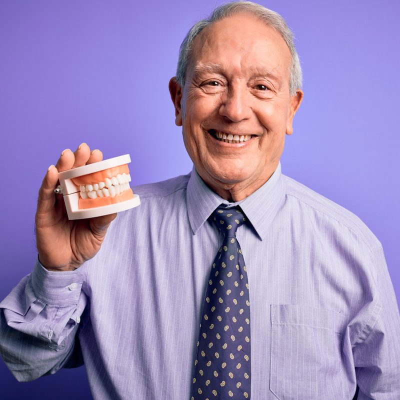 Grey haired senior man holding orthodontic prosthesis denture over purple background with a happy face standing and smiling with a confident smile showing teeth