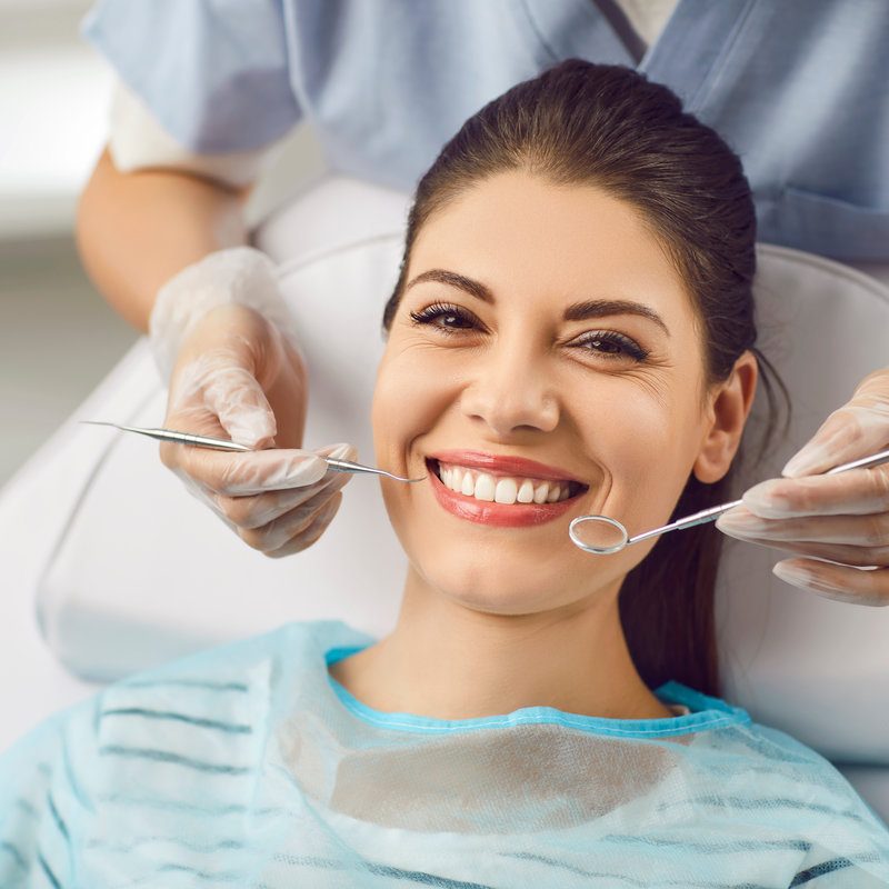 Portrait of a smiling patient with dentist doctor is checking teeth with a dental instrument. Dental care provided during the medical visit, regular dentistry check up for maintaining oral health.