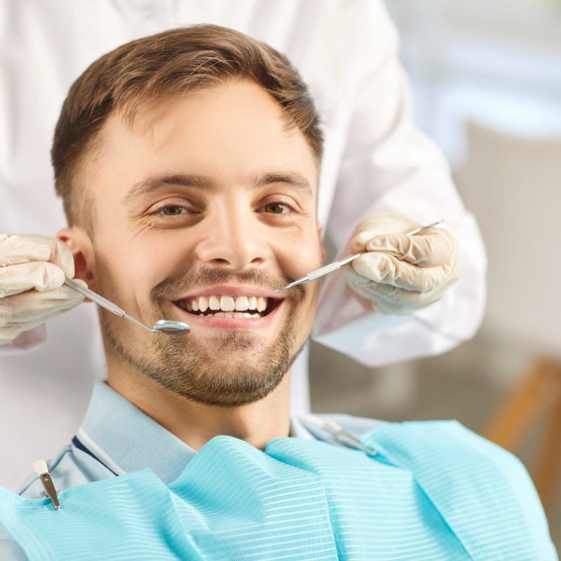 Portrait of male patient looking cheerful at camera in modern dentistry clinic during medical check up in medical center. Dentist examining young man teeth with dental tools. Dental health care.