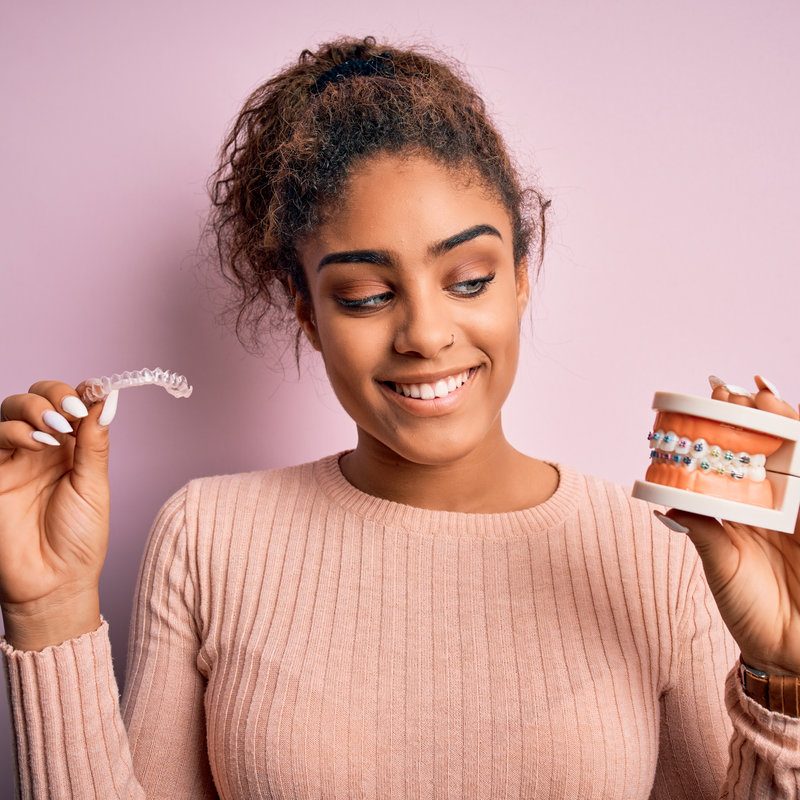 Young african american woman smiling happy holding professional orthodontic denture with metal braces and removable invisible aligner. Comparation of two dental straighten treatments